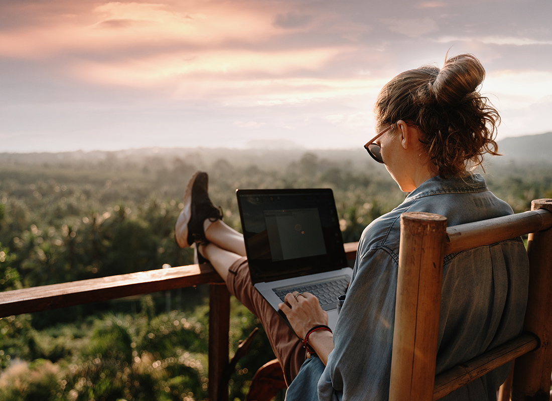 Blog - Young Adventurous Business Woman Sitting on a Deck Overlooking Moutains and Trees While Looking at Her Open Laptop on a Beautiful Day